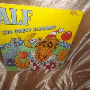May include: A yellow book cover titled ALF THE GREAT ALFONSO featuring a cartoon illustration of the character Alf wearing a clown costume with a red and white hat, a banana, a red apple, an orange, and a small gray seal on a blue base.