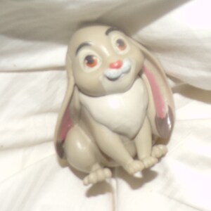May include: A small, brown rabbit figurine with pink ears and large, brown eyes. The rabbit is sitting with its paws tucked under its body.