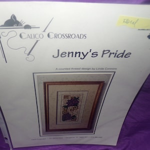Op de afbeelding: Een geteld draad kruissteekpatroon van Calico Crossroads genaamd "Jenny's Pride", ontworpen door Linda Connors. Het patroon toont een paarse bloem met groene bladeren in een ingelijst beeld.