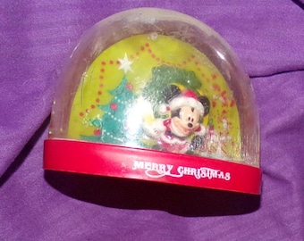 May include: A clear plastic snow globe with a red base. The globe features a cartoon image of Mickey Mouse in a Santa hat, standing in front of a green Christmas tree. The base has the words "Merry Christmas" printed on it.