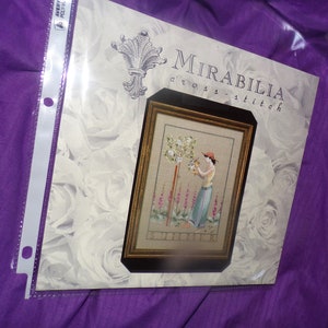 May include: A cross-stitch pattern featuring a woman in a red hat and blue dress tending to a white rose bush. The pattern is titled "Summer" and is from the Mirabilia cross-stitch collection.