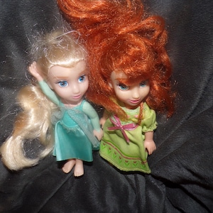 May include: Two small dolls with detailed facial features and flowing hair. One doll has blonde hair and a teal dress, the other has red hair and a green dress. Both dolls are posed on a dark, textured surface.