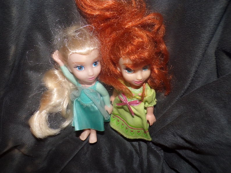 May include: Two small dolls with detailed facial features and flowing hair. One doll has blonde hair and a teal dress, the other has red hair and a green dress. Both dolls are posed on a dark, textured surface.