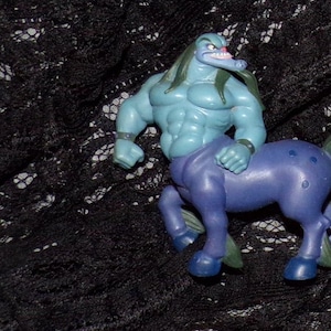 May include: A blue and purple plastic toy figure of a centaur. The figure has a muscular upper body and a horse's lower body. The figure is posed in a running position.