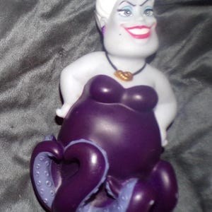 May include: A figurine of Ursula, the sea witch from Disney's The Little Mermaid. The figure has a white head with a spiky hairstyle, a purple body, and tentacles. She wears a black necklace with a gold pendant and has a wide smile.