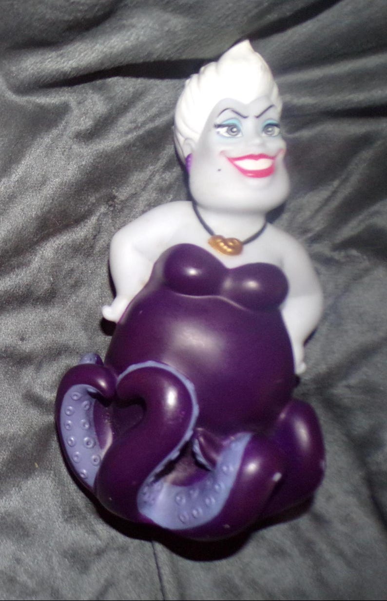 May include: A figurine of Ursula, the sea witch from Disney's The Little Mermaid. The figure has a white head with a spiky hairstyle, a purple body, and tentacles. She wears a black necklace with a gold pendant and has a wide smile.