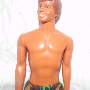 May include: A vintage Ken doll with brown hair and blue eyes. The doll has a tan complexion and is shirtless, wearing patterned shorts with brown, green, and blue designs. The doll is smiling.