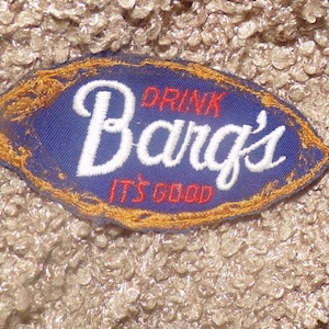 May include: An oval-shaped embroidered patch featuring a blue background with a gold border. The text reads DRINK in red, Barg's in large white script, and IT'S GOOD in red. The patch is placed on a textured, light brown fabric surface.