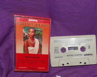 May include: A cassette tape with a red and white label featuring a photo of Whitney Houston. The label reads "Whitney Houston" and "Arista". The cassette tape is labeled "Whitney Houston" and "Arista" and has the song title "How Will I Know" and "Greatest Love Of All".