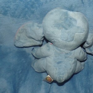 Puede incluir: Juguete de peluche azul de Stitch de Lilo & Stitch. El juguete est&aacute; sentado en una manta azul. El juguete tiene una nariz rosa y orejas interiores rosas.