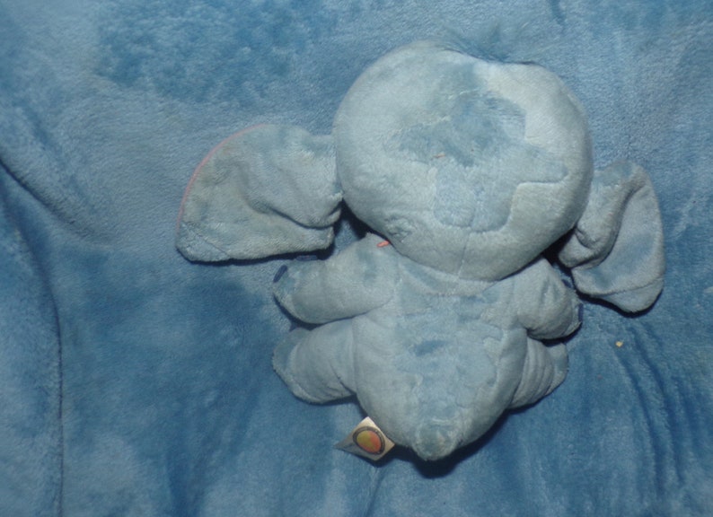 Puede incluir: Juguete de peluche azul de Stitch de Lilo & Stitch. El juguete est&aacute; sentado en una manta azul. El juguete tiene una nariz rosa y orejas interiores rosas.