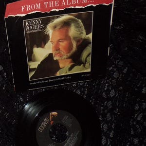 May include: A black and white 45 RPM vinyl record with a black label. The label has the RCA logo and the text "What About Me?" by Kenny Rogers and Kim Carnes. The record is on a black lace surface. The record is from the album "What About Me?" by Kenny Rogers.