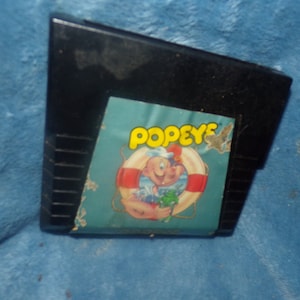 May include: A black video game cartridge featuring a label with the text POPEYE in yellow letters. The label displays a cartoon illustration of Popeye the Sailor inside a red and white life preserver, holding a green object against a blue background.