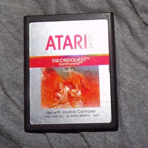 May include: A vintage Atari 2600 video game cartridge for Swordquest: Earthworld. The label features the Atari logo in red, the game title, and a colorful illustration of two figures. The cartridge is black with a silver label.