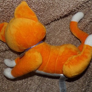 May include: Orange plush cat toy with white paws and tail tip. The cat has a purple collar and is lying down on a brown surface. The toy is made of soft material and has stitched details.