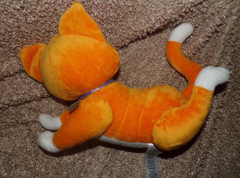 May include: Orange plush cat toy with white paws and tail tip. The cat has a purple collar and is lying down on a brown surface. The toy is made of soft material and has stitched details.