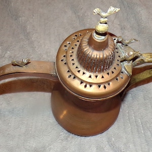 May include: A copper coffee pot with a decorative lid and a bird-shaped handle. The pot has a traditional design and is perfect for brewing coffee or tea.
