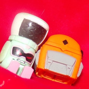 May include: Two small plastic toys, one is green and white with a black visor and the other is orange with a gray front and a black diamond on top.