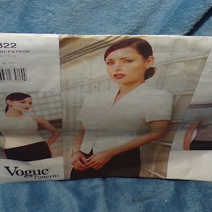 May include: A sewing pattern for a women's blouse, featuring a white blouse with a collar and a white and black striped blouse. The pattern is from Vogue Patterns and is designed by Anne Klein II. The pattern number is 1822.