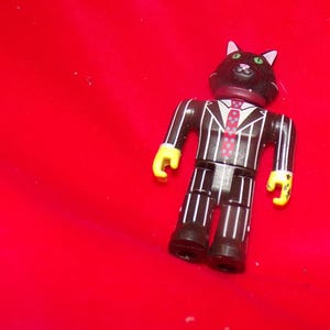 May include: A black and white striped suit with a black cat head and a red and white polka dot tie. The figure has yellow hands and is standing on a red surface.