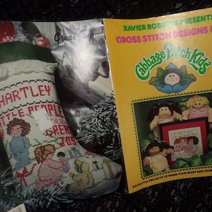 May include: A cross stitch pattern booklet featuring designs for Cabbage Patch Kids. The cover features a yellow background with the Cabbage Patch Kids logo and the text "Xavier Roberts Presents Cross Stitch Designs for Cabbage Patch Kids". The booklet includes a variety of designs, including a stocking with the text "Hartley Little People" and a design featuring a group of Cabbage Patch Kids.