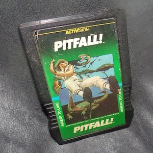 May include: A black Activision Pitfall! video game cartridge. The label features a jungle scene with a man swinging on a vine, a crocodile, and snakes. The game title "PITFALL!" is prominently displayed.