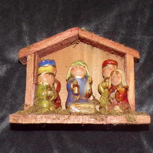 May include: A wooden nativity scene with five ceramic figures. The figures are painted in bright colors, including red, blue, green, yellow, and gold. The figures are arranged in a manger, with a small amount of moss around them.