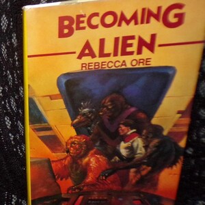 May include: A paperback book titled "Becoming Alien" by Rebecca Ore. The cover features a yellow background with red text and an illustration of several alien figures and a human in a spaceship setting.