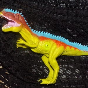 May include: A yellow, orange, and blue toy dinosaur with an open mouth, revealing pink. The dinosaur has a spiky blue ridge down its back. The toy is on a black, textured surface.