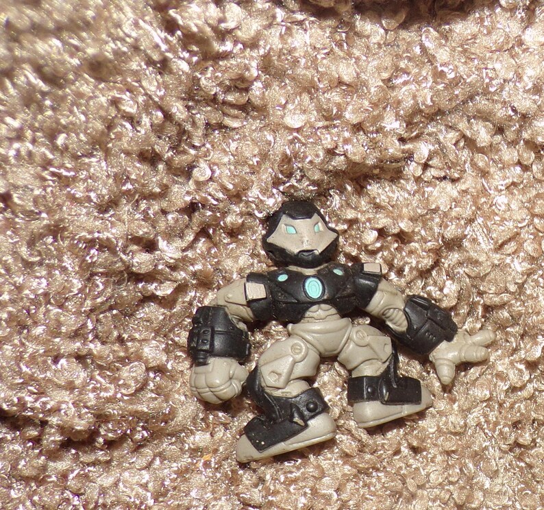 K&ouml;nnte beinhalten: Eine kleine Actionfigur eines roboterartigen Charakters. Die Figur ist haupts&auml;chlich schwarz, grau und beige, mit einem hellblauen kreisf&ouml;rmigen Design auf der Brust. Die Figur hat einen helm&auml;hnlichen Kopf und bewegliche Gliedma&szlig;en.