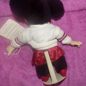 Puede incluir: Un juguete de peluche de un personaje blanco y negro que lleva una camisa blanca con ribetes rojos, una falda roja y polainas negras. El juguete est&aacute; de pie sobre un soporte blanco.