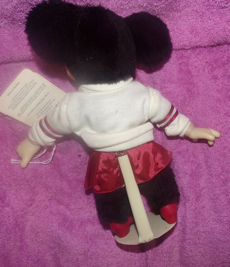 Puede incluir: Un juguete de peluche de un personaje blanco y negro que lleva una camisa blanca con ribetes rojos, una falda roja y polainas negras. El juguete est&aacute; de pie sobre un soporte blanco.
