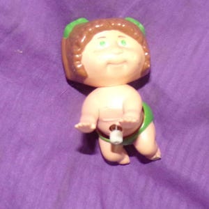 May include: A wind-up toy of a Cabbage Patch Kid doll with brown hair, green bows, and a green diaper. The doll is wearing a green diaper and has a wind-up key on its back.