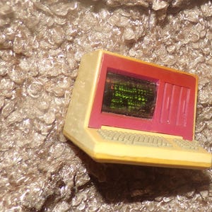 May include: A miniature beige and magenta plastic computer toy featuring a screen with printed text that reads Barbie, Mattel, and Skipper. The device includes a molded keyboard and a vertical panel on the right side of the screen frame.