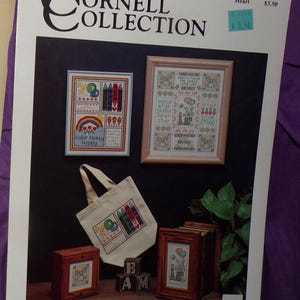 May include: The Cornell Collection catalog cover featuring cross-stitch patterns. The cover includes framed cross-stitch samplers, a tote bag, and small wooden framed pieces. The text "color today happy" is visible on one sampler.