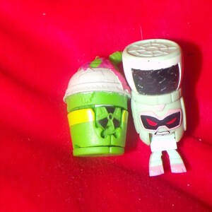 May include: Two green and white toy figures. One figure is a robot with red eyes and a black and white design. The other figure is a green and white cup with a yellow stripe and a black and white design.