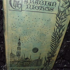May include: A vintage book cover with a green background and black illustrations. The title "The Arabian Nights" is printed in black lettering. The cover features a crescent moon, a minaret, and a palm tree.
