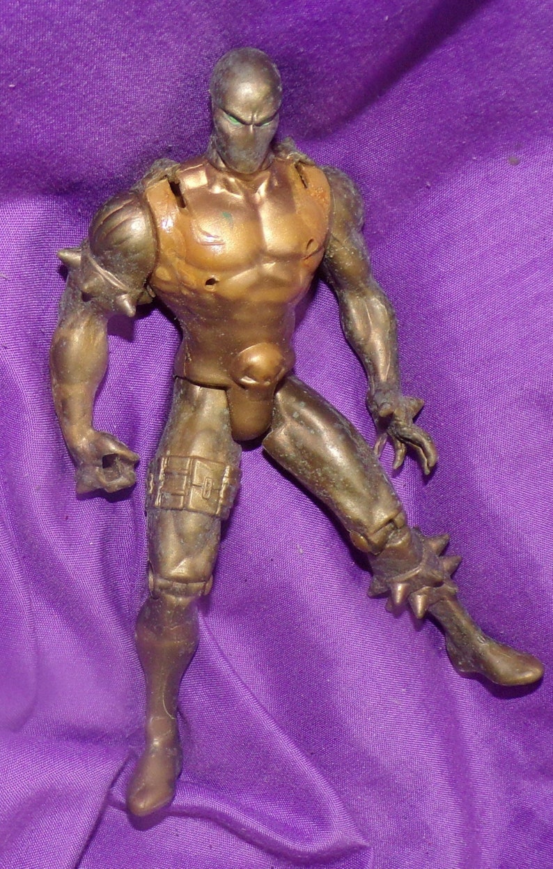 May include: A gold-colored action figure with a green face mask and spiky armor. The figure is posed with one leg raised and one arm extended.