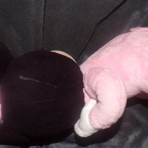 K&ouml;nnte beinhalten: Ein Pl&uuml;schtier in Form einer Figur, mit einem gro&szlig;en schwarzen Kopf mit runden Ohren und einem rosa K&ouml;rper. Das Spielzeug hat wei&szlig;e Handschuhe und ein Etikett an seinem Fu&szlig;. Das Spielzeug liegt auf einer dunklen Oberfl&auml;che.