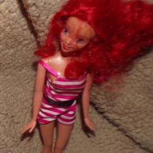 May include: A red-haired doll wearing a pink and white striped swimsuit with a black belt.
