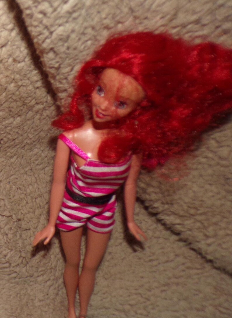 May include: A red-haired doll wearing a pink and white striped swimsuit with a black belt.