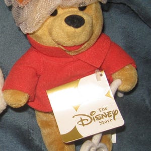 May include: A Winnie the Pooh plush toy wearing a red shirt and an orange hat with a white veil. The Disney Store tag is attached to the toy. The plush toy is a classic character from the Disney franchise.