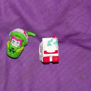 May include: Two small plastic toys, one green and one white. The green toy is shaped like a milk carton with a pink and green face and a green straw. The white toy is shaped like a milk carton with a red and white design and the text "05-5487 90721".