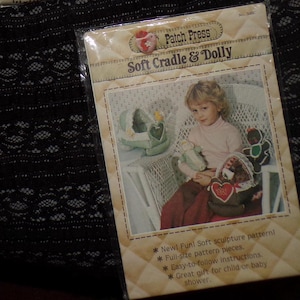 Könnte beinhalten: Eine Verpackung für ein Soft-Skulpturenmuster "Soft Cradle & Dolly". Die Verpackung zeigt ein Kind, das eine Puppe und andere weiche Skulpturen hält. Der Text lautet "New! Fun! Soft sculpture pattern!" und "Easy-to-follow instructions."