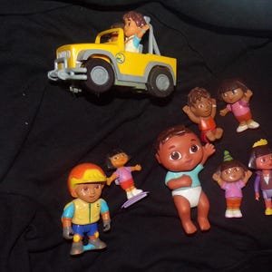 May include: A collection of children's toy figures and a yellow jeep. The figures include Dora the Explorer and Diego, with various outfits and poses. The jeep has a figure in the driver's seat.