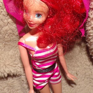 May include: A red-haired doll wearing a pink and white striped swimsuit with a black belt. The doll has blue eyes and is wearing a pink scarf.