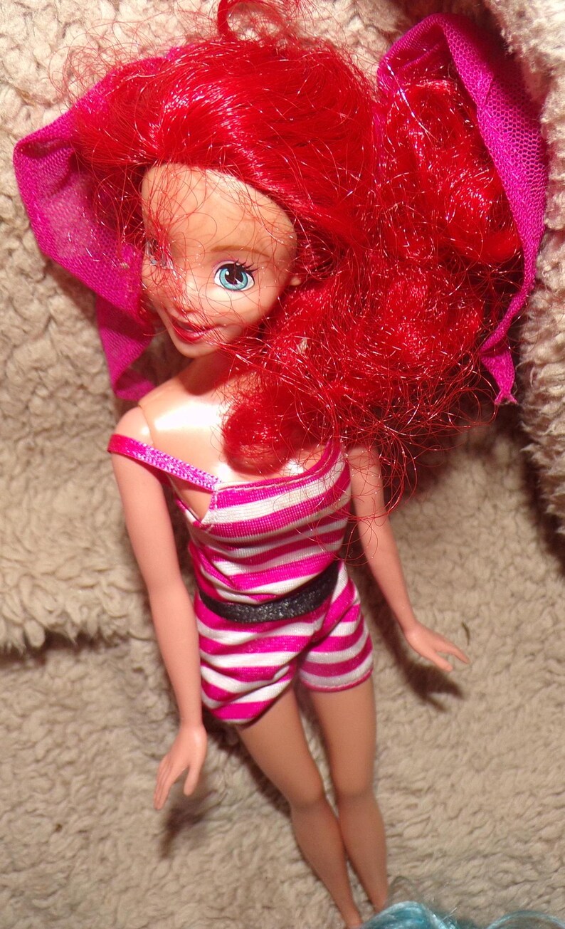 May include: A red-haired doll wearing a pink and white striped swimsuit with a black belt. The doll has blue eyes and is wearing a pink scarf.