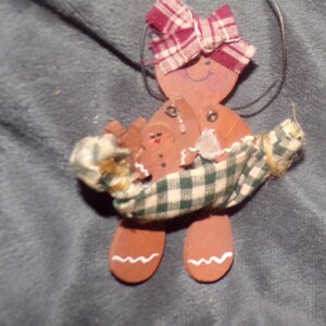 May include: Wooden gingerbread man Christmas ornament with a red and white plaid bow and a green and white plaid blanket. The ornament has a wire loop for hanging and is decorated with a smaller gingerbread man.
