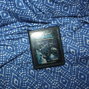 May include: A black Atari game cartridge for the game Asteroids. The cartridge has the game title and a graphic of a spaceship battle. The cartridge is labeled "Program Asteroids" and "Use with Joystick Controllers."