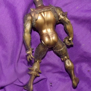 May include: A gold-colored plastic action figure with a spiky helmet and armor. The figure is posed with its arms raised and its legs spread apart. The figure is made of plastic and has a matte finish. The figure is standing on a purple surface.